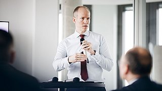 Foto, Mann in weißem Hemd und roter Krawatte hält Mikrofon und spricht vor Publikum in einem modernen Raum