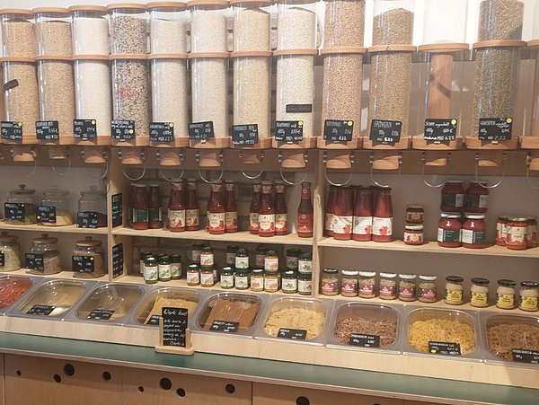 Foto, Regal mit mehreren großen Spenderbehältern für Getreide und Hülsenfrüchte sowie darunter stehenden Gläsern und Schalen mit verschiedenen Lebensmitteln in einem Laden