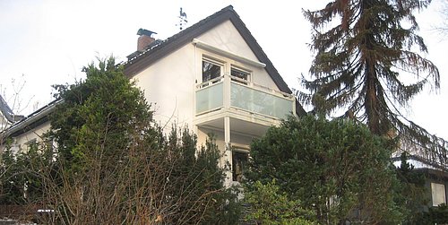 Foto, Außenansicht eines weißen Fertighauses aus den 70er Jahren im Winter mit Balkon und umgeben von Nadelbäumen und Sträuchern