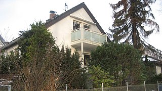 Foto, Außenansicht eines weißen Fertighauses aus den 70er Jahren im Winter mit Balkon und umgeben von Nadelbäumen und Sträuchern