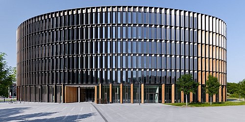 Foto, Außenansicht des Rathauses in Freiburg, einem mehrstöckigen, runden Gebäudes mit einer von Holzpanele und Photovoltaikmodulen durchbrochenen Glasfassade.