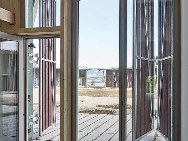 Foto, Blick durch große Fenster und Glastüren eines modernen Holzhauses auf eine Wohnsiedlung mit weiteren Gebäuden und offener Landschaft