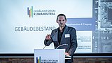 Foto, Person steht am Rednerpult mit Mikrofon beim Gebäudeforum Klimaneutral, Präsentation im Hintergrund