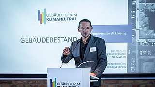 Foto, Person steht am Rednerpult mit Mikrofon beim Gebäudeforum Klimaneutral, Präsentation im Hintergrund