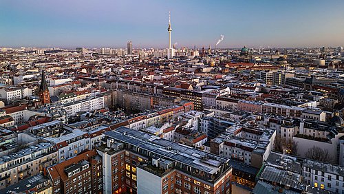 Foto, Luftaufnahme einer Stadt mit dicht bebauten Wohngebäuden und einem hohen Fernsehturm im Hintergrund