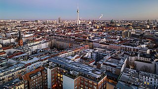 Foto, Luftaufnahme einer Stadt mit dicht bebauten Wohngebäuden und einem hohen Fernsehturm im Hintergrund