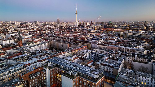 Foto, Luftaufnahme einer Stadt mit dicht bebauten Wohngebäuden und einem hohen Fernsehturm im Hintergrund