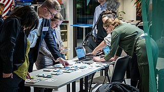 Foto, mehrere Personen stehen an einem Tisch mit ausgebreiteten Flyern und Broschüren, eine Person arbeitet an einem Laptop.