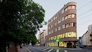 Foto, Ansicht von der Straße eines mehrstöckigen, spitz zulaufenden Gebäudes mit Holzfassade. Links im Bild steht ein Baum.