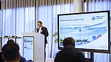 Foto, ein Redner präsentiert auf einer Konferenz, im Hintergrund sind auf einem großen Bildschirm Veranstaltungshinweise zu sehen.