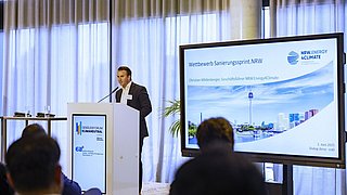 Foto, ein Redner präsentiert auf einer Konferenz, im Hintergrund sind auf einem großen Bildschirm Veranstaltungshinweise zu sehen.
