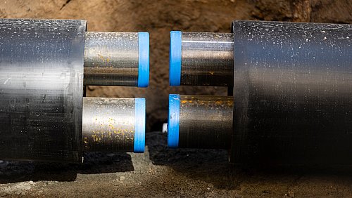Foto, zwei Metallrohre mit blauen Gummienden sind in einem Graben verbunden, der von Erde und Kies umgeben ist.