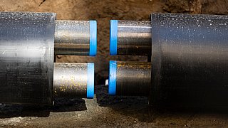 Foto, zwei Metallrohre mit blauen Gummienden sind in einem Graben verbunden, der von Erde und Kies umgeben ist.