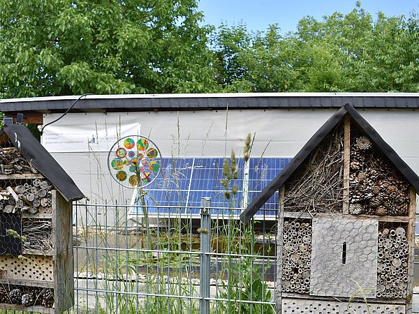 Foto, Zwei Insektenhotels im Klimagarten am Zaun hängend, dahinter Solarmodule am Grünen Klassenzimmer