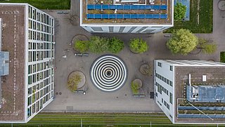 Foto, Luftaufnahme dreierGebäude mit PV-Modulen auf dem Dach, die sich um einen kreisrunden Brunnen gruppieren.