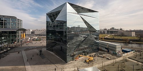 Foto, Sicht von oben auf spiegelndes, quadratisches Gebäude. Vor dem Gebäude befindet sich eine Baustelle und Baustellenfahrzeuge.