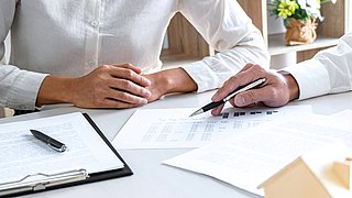 Foto, zwei Personen in weißen Hemden sitzen an einem Tisch, eine zeigt mit einem Stift auf ein Blatt mit Diagrammen und Tabellen.