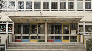 Foto, Eingangsbereich einer Schule mit farbigen Türen, Treppen und bunt bemalten Fenstern.