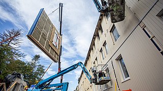 Foto, Bauarbeiten an einer Hausfassade. Mit einer Hebebühne wird ein Fassadenelement zum Haus befördert.