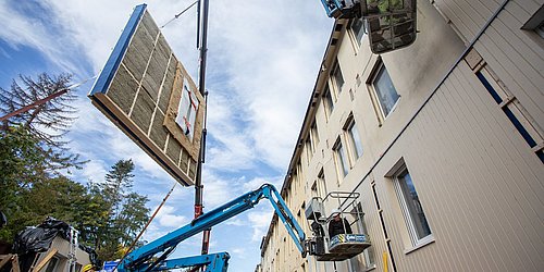 Foto, Bauarbeiten an einer Hausfassade. Mit einer Hebebühne wird ein Fassadenelement zum Haus befördert.