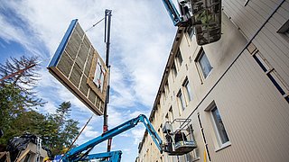 Foto, Bauarbeiten an einer Hausfassade. Mit einer Hebebühne wird ein Fassadenelement zum Haus befördert.