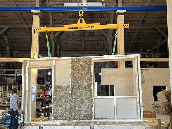 Foto, In einer Werkhalle hängt ein gelber Kranbalken mit der Aufschrift 'MITARI WLL 4000 KG' an Seilen, darunter ein Holzrahmen mit zwei mit Stroh gefüllten Flächen, im Hintergrund stehen zwei Personen und Regale mit Werkzeugen.