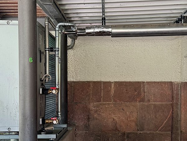 Foto, Eine Wärmepumpe mit metallischen Rohrleitungen ist an einer Hauswand montiert. Die Rohre verlaufen waagerecht entlang der Wand und sind teilweise mit Isoliermaterial ummantelt. Das Gerät steht auf einem Metallgestell, und an der Wand ist eine Außenleuchte befestigt. Die Wand besteht aus verputztem Mauerwerk und roten Sandsteinquadern. Das Dach besteht aus Wellblech.