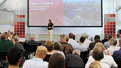 Foto, Vortragende Person spricht vor Publikum. Im Hintergrund ist eine Großleinwand mit dem Titel der Konferenz und einem Bild von Hamburg zu sehen.