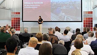 Foto, Vortragende Person spricht vor Publikum. Im Hintergrund ist eine Großleinwand mit dem Titel der Konferenz und einem Bild von Hamburg zu sehen.
