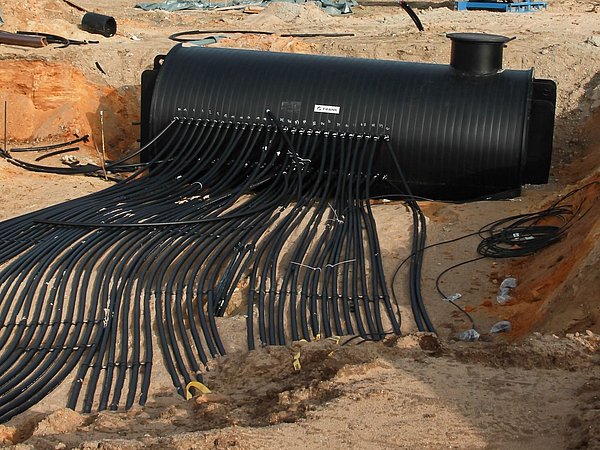 Foto, Schacht mit zahlreichen schwarzen Leitungen auf einem Erdsondenfeld vor Industriegebäuden und einem roten Backsteinhaus