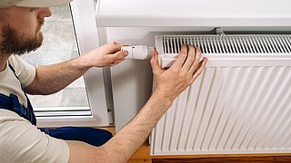 Foto, Handwerker stellt Thermostatventil an einem Heizkörper in einem Raum mit Holzboden ein.