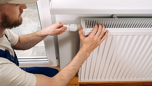 Foto, Handwerker stellt Thermostatventil an einem Heizkörper in einem Raum mit Holzboden ein.