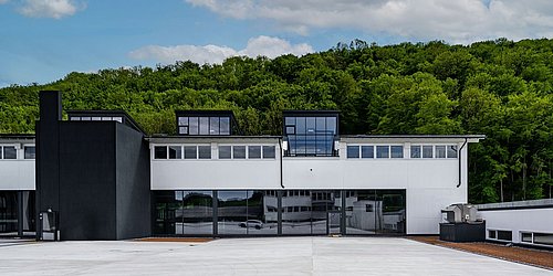 Foto, Handelszentrum mit weißer Fassade und großen Fenstern vor bewaldetem Hügel unter blauem Himmel