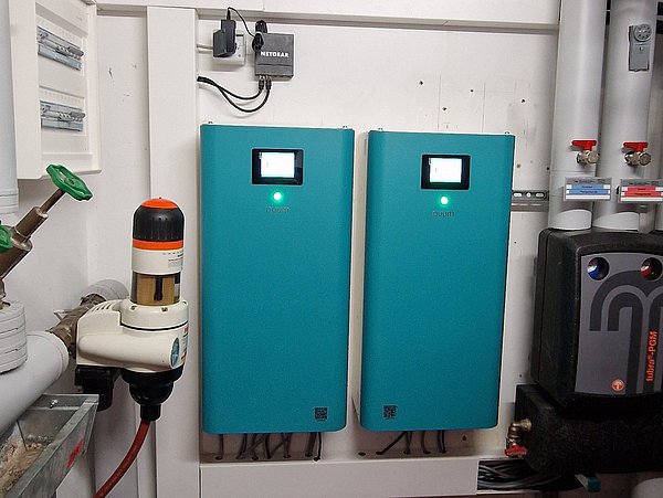 Foto, Inneneinheit der Wärmepumpenkaskade, Technikraum mit zwei blauen Steuergeräten an der Wand, mehreren Rohrleitungen und Ventilen