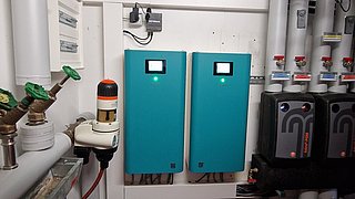 Foto, Inneneinheit der Wärmepumpenkaskade, Technikraum mit zwei blauen Steuergeräten an der Wand, mehreren Rohrleitungen und Ventilen