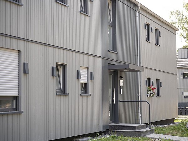 Foto, Mehrfamilienhaus mit grauer Holzverkleidung, mehreren Fenstern und einem Eingang mit Treppe und Handlauf