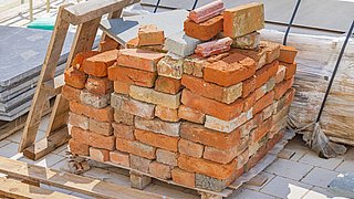 Foto, auf einem Holzpalettenstapel liegen unregelmäßig gestapelte Ziegelsteine und einige Betonplatten im Hintergrund.