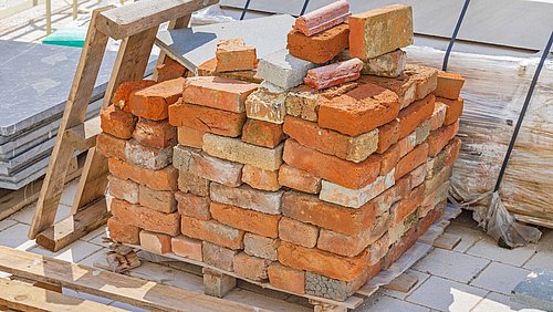 Foto, auf einem Holzpalettenstapel liegen unregelmäßig gestapelte Ziegelsteine und einige Betonplatten im Hintergrund.
