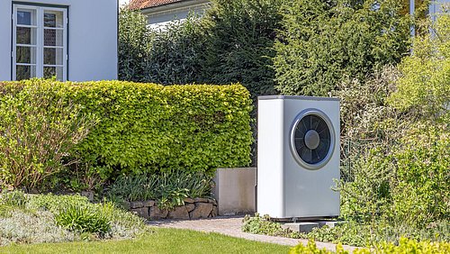 Foto, Weißes, rechteckiges Außengerät einer Wärmepumpe mit rundem Lüfter vor einem Haus mit rotem Ziegeldach und grüner Hecke.