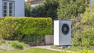 Foto, Weißes, rechteckiges Außengerät einer Wärmepumpe mit rundem Lüfter vor einem Haus mit rotem Ziegeldach und grüner Hecke.