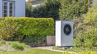 Foto, Weißes, rechteckiges Außengerät einer Wärmepumpe mit rundem Lüfter vor einem Haus mit rotem Ziegeldach und grüner Hecke.