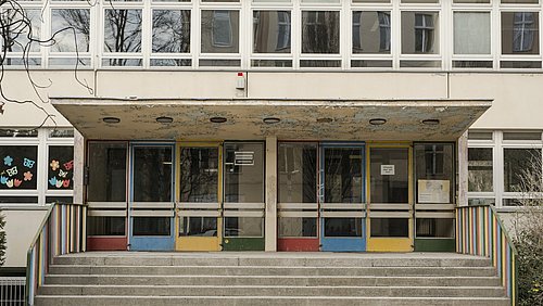 Foto, Eingangsbereich einer Schule mit farbigen Türen, Treppen und bunt bemalten Fenstern.