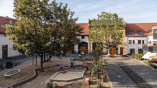 Foto, Spielplatz mit Sandkasten und Klettergerüst zwischen Bäumen in einem Innenhof mit Wohnhäusern und roten Dächern