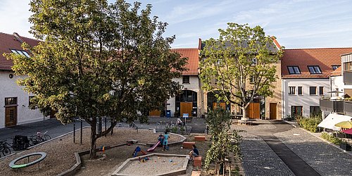 Foto, Spielplatz mit Sandkasten und Klettergerüst zwischen Bäumen in einem Innenhof mit Wohnhäusern und roten Dächern