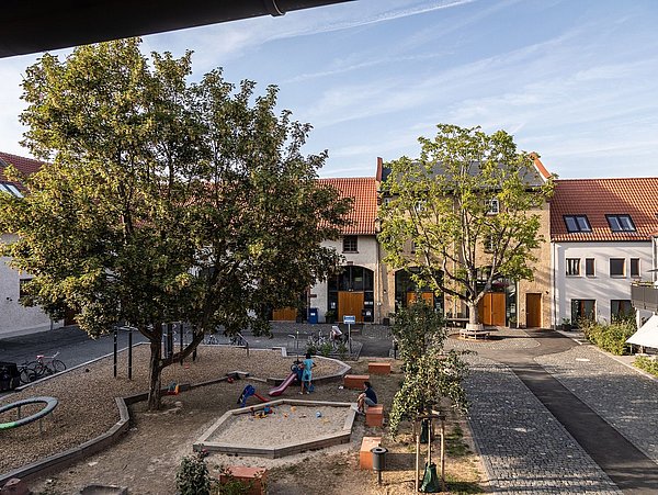Foto, Spielplatz mit Sandkasten und Klettergerüst zwischen Bäumen in einem Innenhof mit Wohnhäusern und roten Dächern