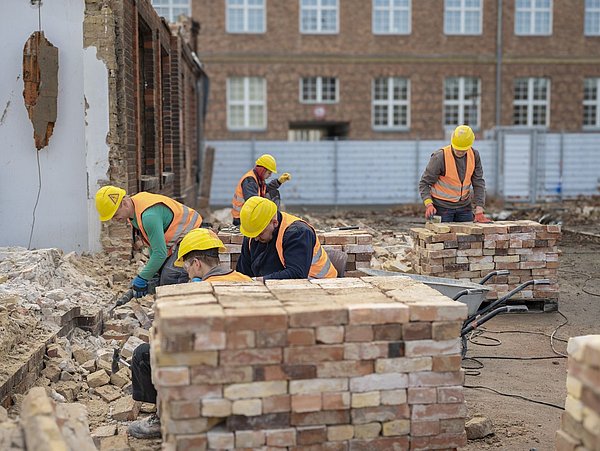 Foto, Fünf Bauarbeiter mit gelben Helmen und orangen Warnwesten mauern Ziegelwände auf einer Baustelle mit altem Backsteingebäude im Hintergrund.