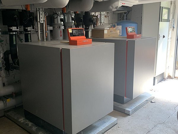 Foto, Zwei große graue Wärmepumpen mit orangefarbenen Steuerungen in einem Technikraum mit weißen Rohrleitungen