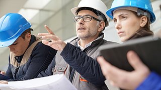 Foto, Nahaufnahme einer Gruppe von Menschen auf einer Baustelle, die einen Plan studieren. Eine Person deutet mit dem Finger aud etwas ausßerhalb des Bildauschnittes.