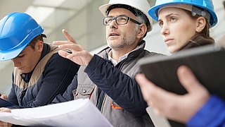 Foto, Nahaufnahme einer Gruppe von Menschen auf einer Baustelle, die einen Plan studieren. Eine Person deutet mit dem Finger aud etwas ausßerhalb des Bildauschnittes.