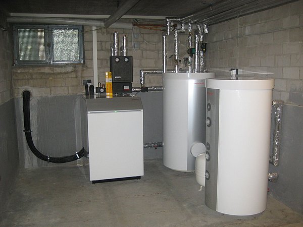 Foto, Heizungsanlage mit zwei weißen Warmwasserspeichern und verrohrtem Heizkessel in einem Kellerraum mit Betonwänden und kleinem Fenster